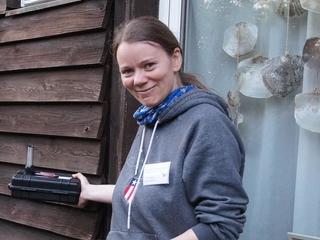 Eine Frau steht vor einer Holzfassade und hält einen Fledermausdetektor an die Fassade.