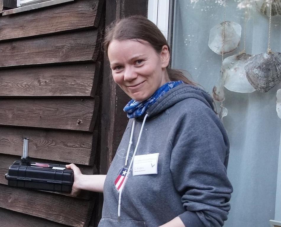 Eine Frau steht vor einer Holzfassade und hält einen Fledermausdetektor an die Fassade.