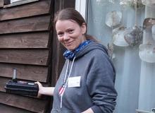 Eine Frau steht vor einer Holzfassade und hält einen Fledermausdetektor an die Fassade.