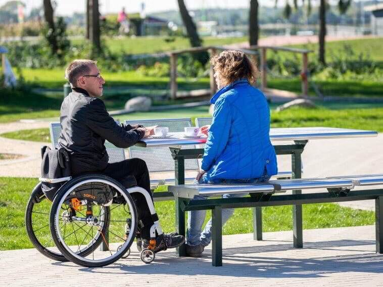 Eine Frau und ein Mann im Rollstuhl picknicken an einem unterfahrbaren Picknickplatz im Grünen.