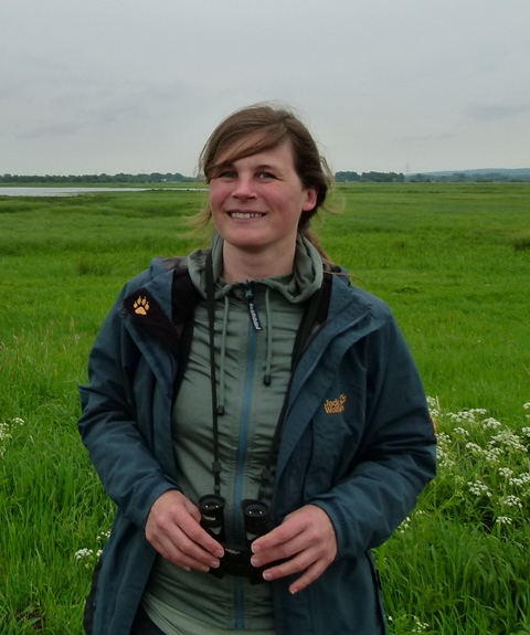 Eine Frau steht auf einer Wiese und hat ein Fernglas um den Hals. Sie lächelt freundlich in die Kamera.