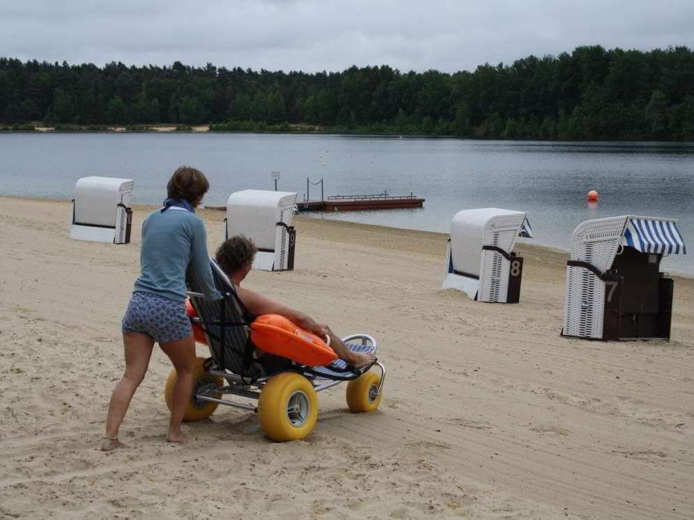 Eine Frau schiebt einen Mann im Strandrollstuhl über den Sand, ans Wasser.