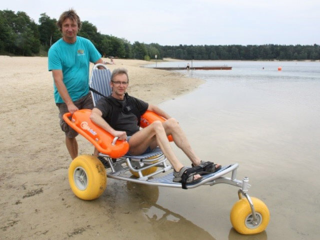 ein stehender Mann und ein sitzender Mann im Strand-Rollstuhl am Seeufer