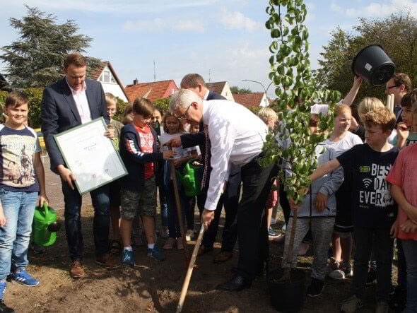 Lehrer und Schüler pflanzen einen Baum. Ein Mann hält eine gerahmte Urkunde in der Hand.