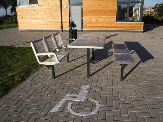 Eine Sitzmöglichkeit auf gepflastertem Untergrund. Davor eine weiße Bodenmarkierung, ein Rollstuhl-Piktogramm.